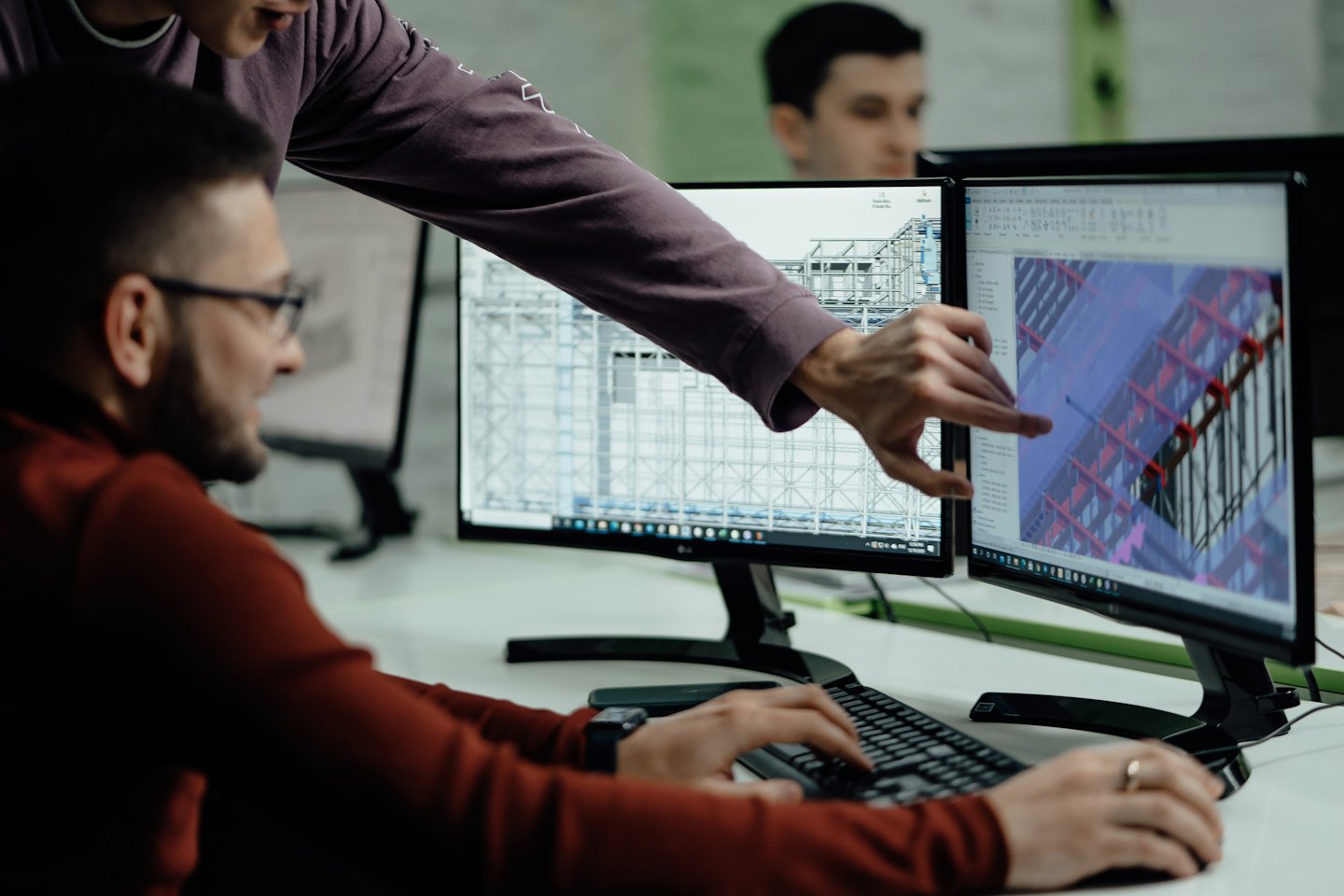 Man points at 3D design on dual monitors while colleague types.